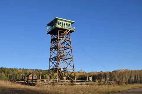 A tall wooden fire lookout tower stands in a clearing surrounded by trees under a clear blue sky.