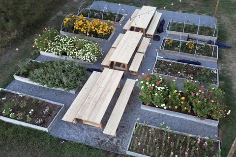 An overhead view shows raised garden beds and wooden picnic tables on gravel.