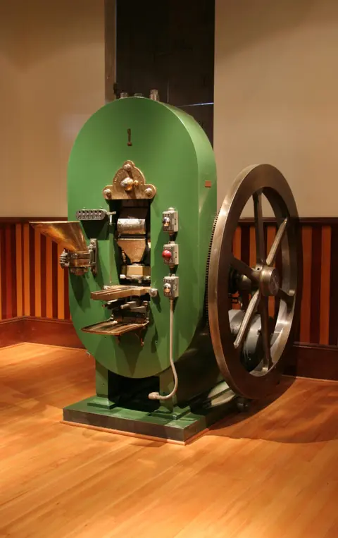 A vintage green and metal industrial machine stands inside a room on a wooden floor.