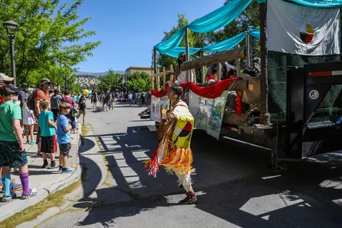 A person dances in traditional regalia alongside a parade float while spectators watch from the street.