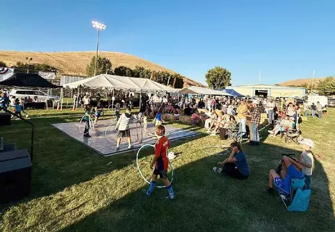 A crowd gathers at an outdoor event with tents and people playing with hula hoops on a platform.
