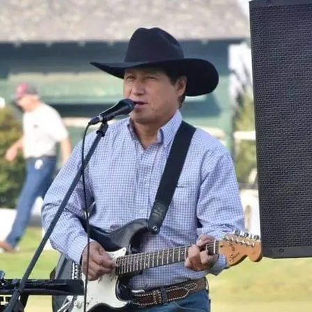 A man wearing a cowboy hat plays an electric guitar and sings into a microphone outdoors.