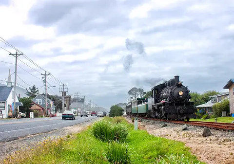 A steam locomotive pulls passenger cars along a track beside a road with cars.