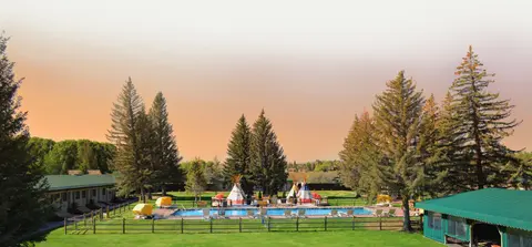 An outdoor swimming pool surrounded by green lawn, trees, and teepees under a warm sky.