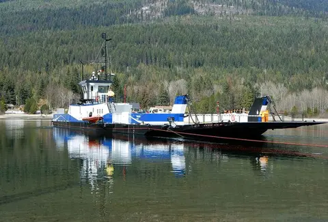 A blue and white cable ferry is on a calm body of water with a forested mountain background.