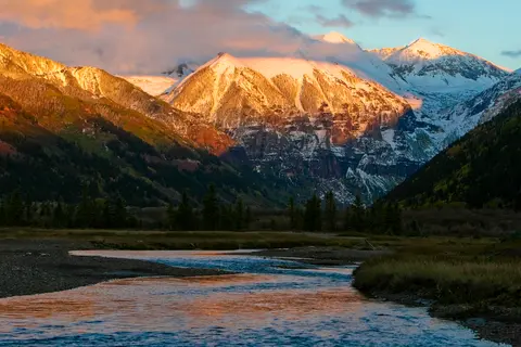 A winding river flows through a valley with snow-capped mountains illuminated by the sun.
