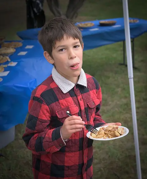A young boy eats pie outdoors near tables.