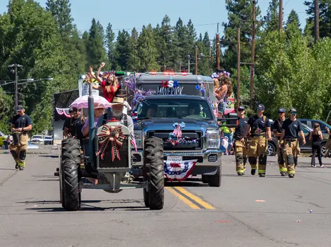 A vintage tractor and decorated truck are driven down a street in a parade.