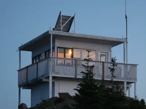 A fire lookout tower stands atop a mountain against a clear blue sky.