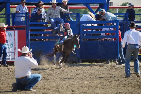 A child rides a horse out of a blue bucking chute at a rodeo.