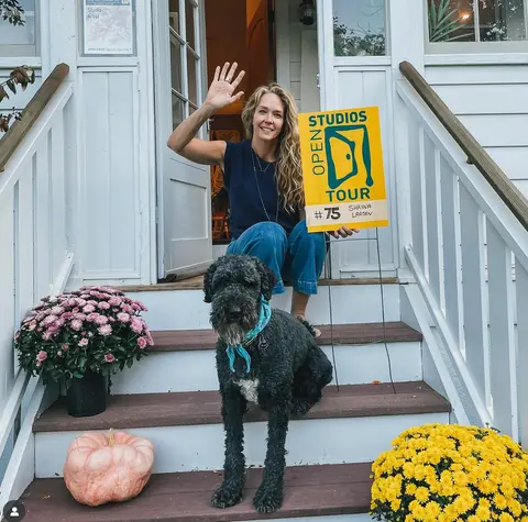 A woman waves while sitting on front steps next to her dog and a tour sign.