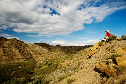 A person looks out over a vast badlands landscape with layered hills and a cloudy sky.