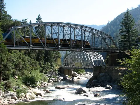 A train crosses a steel bridge over a rushing river in a mountainous landscape.