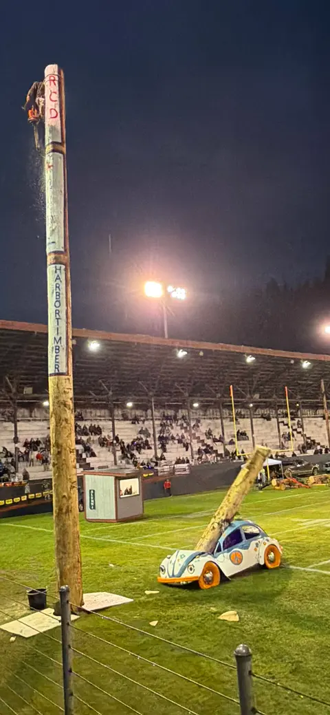 A person cutting the top of a tall wooden pole at an outdoor stadium event at night.