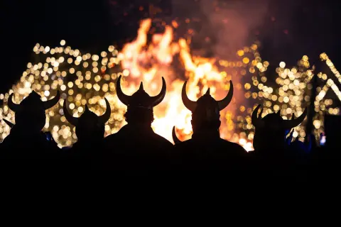 Silhouettes of people wearing horned helmets watch a large bonfire.