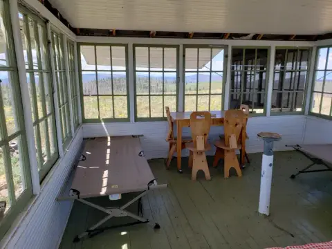 A rustic interior room with large windows, cots, a table, and chairs overlooking a landscape.