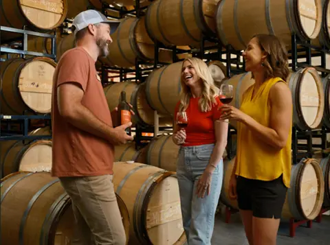 Three people holding wine glasses stand in a cellar surrounded by stacked wine barrels.