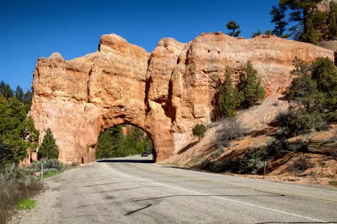 A paved road passes through a natural rock tunnel under a red rock formation.