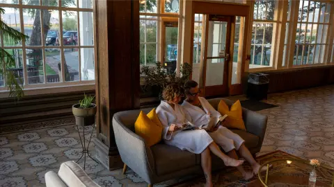 Two people wearing bathrobes sit on a couch inside a sunlit room.