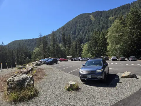 A parking lot sits below a heavily forested mountain under a clear blue sky.