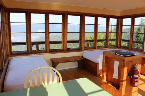 The interior of a fire lookout cabin with panoramic windows and two beds.