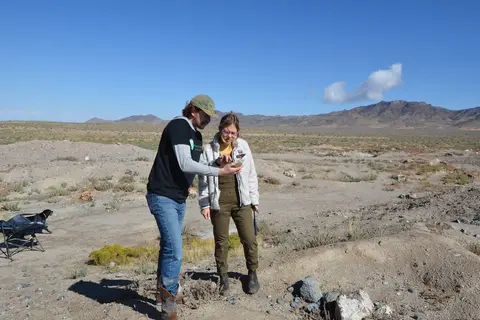 Two people examine an object in an arid desert landscape.