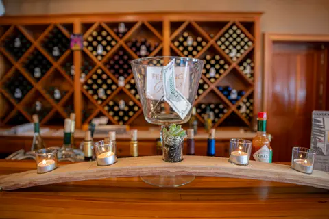 A wooden bar features lit candles in glass holders and a tip jar against a backdrop of wine racks.