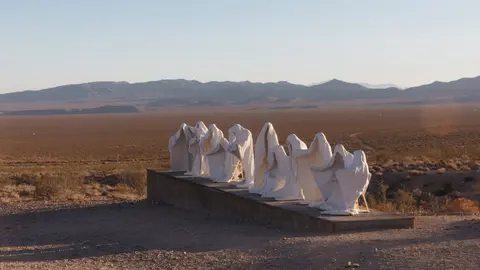 A row of white ghostly sculptures stands on a wooden platform in a desert landscape with mountains in the background.