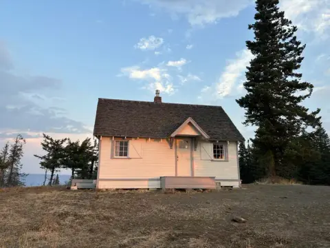 A small cabin stands outdoors surrounded by trees under a cloudy sky.