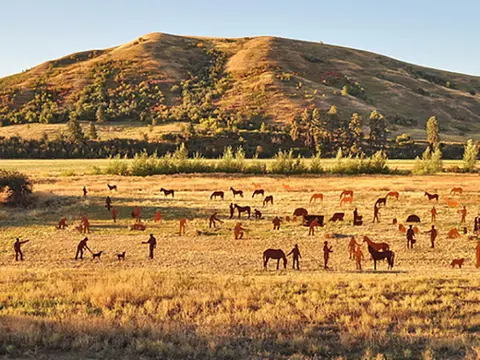 A landscape photograph showing metal silhouette sculptures of people and horses in a field before a grassy hill.