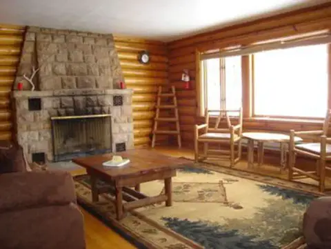 A rustic living room with a stone fireplace, log walls, and large windows.