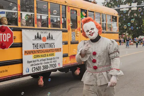 A person in a clown costume stands in front of a decorated yellow bus with bubbles floating in the air.