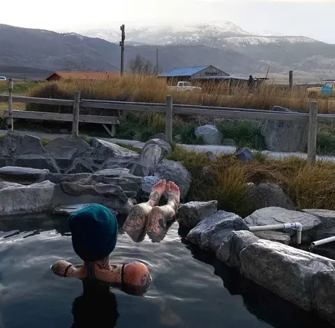 A person relaxing in an outdoor rock hot spring with mountains in the background.