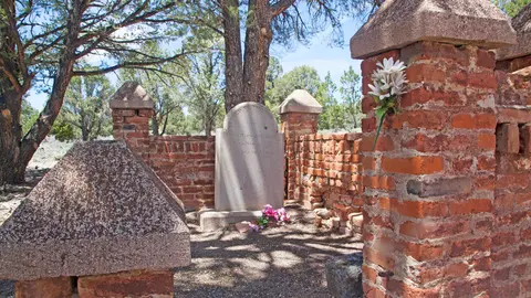 An old gravestone enclosed by a brick wall with stone pillars under trees.