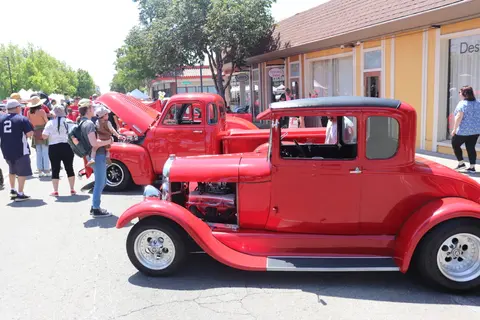 People walk past vintage red cars parked along a street with shops.
