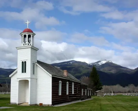 An old white church with a steeple stands in a grassy field with mountains in the background.