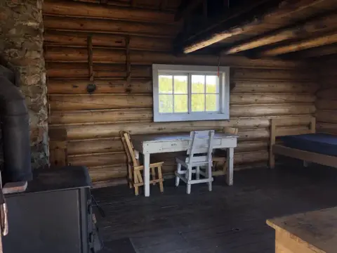 The interior of a rustic log cabin showing a wooden table, chairs, a stove, and a bed.