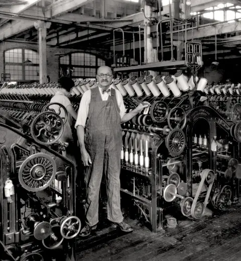 A man in overalls stands by textile machinery in a factory.