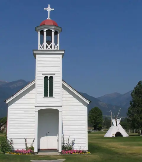 A white wooden church with a bell tower stands in a field with mountains and a teepee in the background.