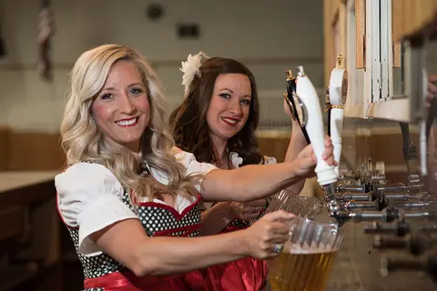 Two women in traditional dirndls pour beer from taps into pitchers.