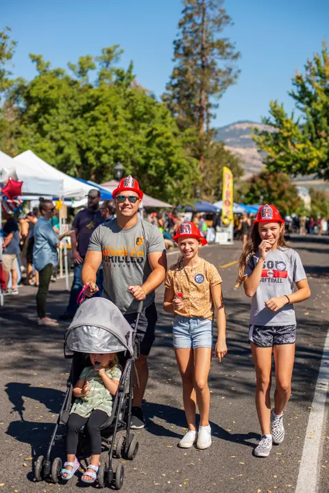 A family walks together on a street during an outdoor event, wearing toy fire helmets.