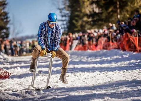 A person riding a makeshift sled down a snowy slope in front of a crowd.