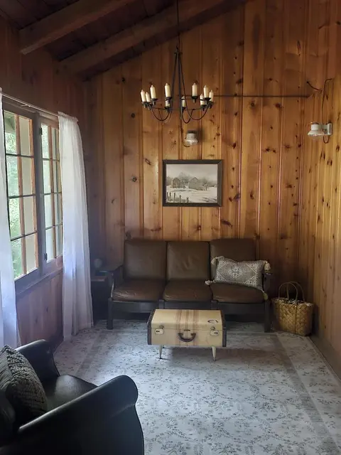 A cozy wood-paneled room features a leather sofa, a chandelier, and a large window with white curtains.