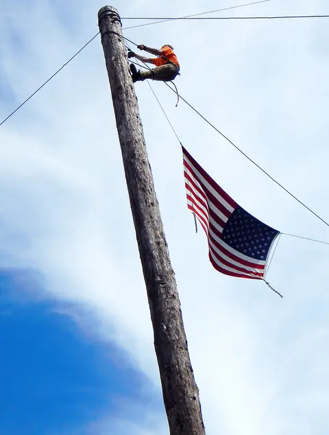 A worker climbs a wooden pole to hang an American flag.