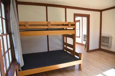 An interior view of a room with a wooden bunk bed, a window on the left, and a doorway to another room.