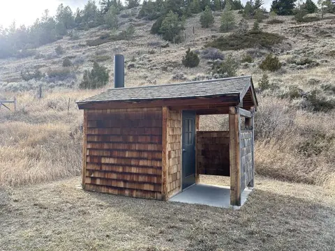 A small wooden outhouse building sits in a grassy area with a hillside behind it.