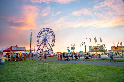 A carnival with a Ferris wheel and food stalls under a sunset sky.