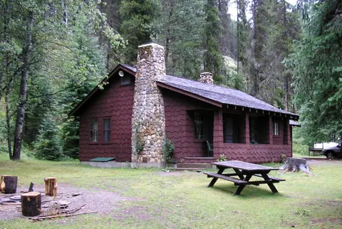 A brown wooden cabin with a large stone chimney sits in a forest clearing.
