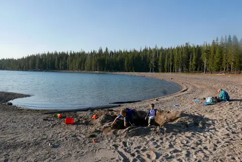 Children play in the sand on a lakeshore with a forest in the background.