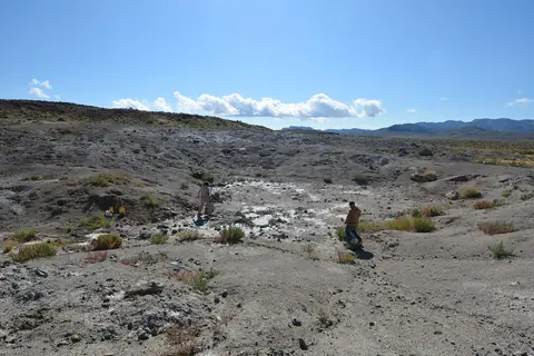 Two people walk across a dry landscape with sparse vegetation under a clear blue sky.